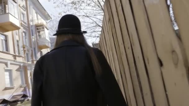 Vue arrière d'une jeune grande fille en veste en cuir noir et un chapeau marchant le long de la clôture en bois dans la rue. L'action. Vue arrière d'une femme marchant dans la rue . — Video