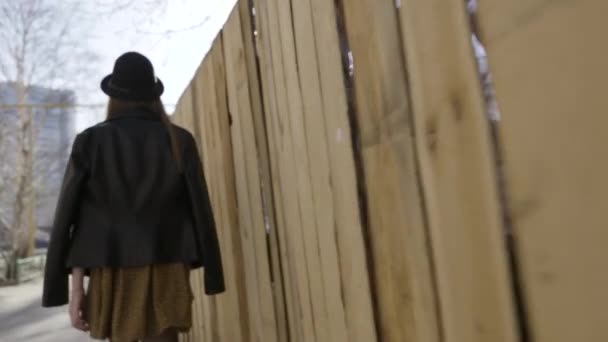 Vue arrière d'une jeune grande fille en veste en cuir noir et un chapeau marchant le long de la clôture en bois dans la rue. L'action. Vue arrière d'une femme marchant dans la rue . — Video