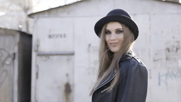 Hezká módní dívka s černým stylovým kloboukem a koženým kabátem pózováním ve staré budově v teplém slunci. Akce. Usměvavý mladý model s kouřovým okem a třepetavou srstí stojící venku. — Stock fotografie