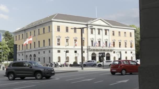Um edifício com muitas bandeiras em uma rua movimentada com carros em movimento, embaixada na Suíça. Acção. Uma rua com veículos de condução perto do edifício municipal bege com bandeiras europeias . — Vídeo de Stock