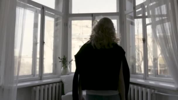Achteraanzicht van blonde in zwarte cardigan op haar schouders beweegt langzaam naar het raam thuis in lichte kamer. Actie. Jonge tiener met krullend haar gaat naar grote Windon in haar appartement. — Stockvideo