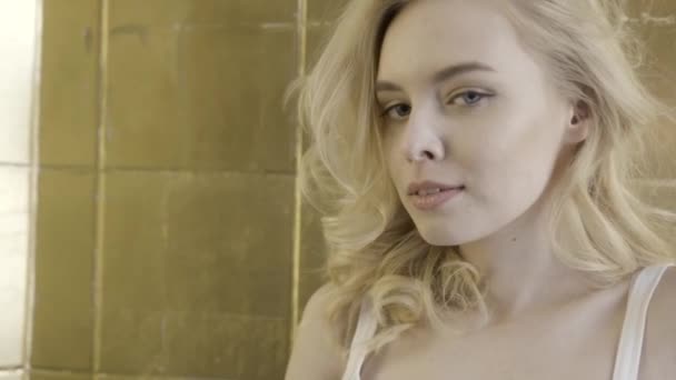 Close up for a beautiful blonde girl face with big blue eyes and curly short hair looking pensive on golden wall background. Action. Young lady posing to the camera and smiling. — Wideo stockowe