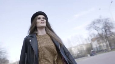 Sokakta çıplak ağaçlar arasında yürüyen bir kadın ve arka planda çocuklu insanlar. Başla. Sonbaharda şehir parkında siyah deri ceket ve şapka giyen genç bir manken kız..