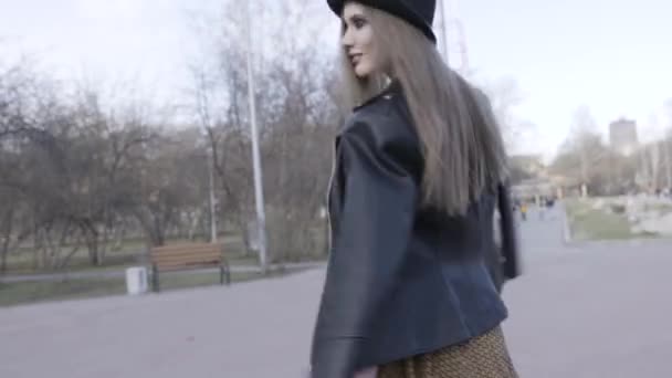 Kvinna promenader längs kala träd i gränden och människor med barn på bakgrunden. Börja. Ung modell flicka i en stadspark på hösten bär svart läder jacka och en hatt. — Stockvideo