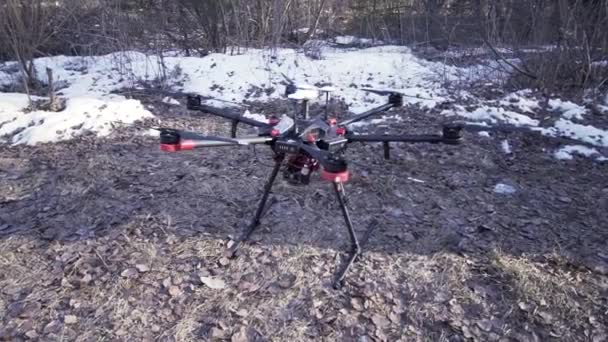 Közel a drón helikopter álló mozdulatlanul a földön a kora tavaszi, videó gyártási koncepció. Csípés. Fekete quadcopter nedves földön, hó és bokrok a háttérben. — Stock videók