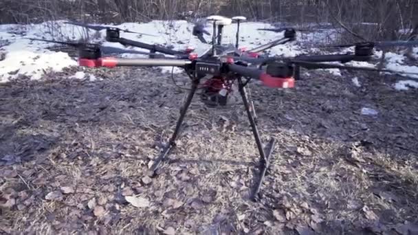 Close up for drone copter standing motionless on the ground in the early spring, video production concept. Clip. Black quadcopter on wet soil with snow and bushes on the background. — Stock Video