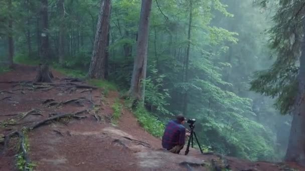 Ekose gömlek ve pantolon derin vadikenarında oturan ve bir sis güzel orman manzara bir resim veya video alarak adam görünümü. Stok görüntüleri. Gizemli ormanın inanılmaz manzarası — Stok video