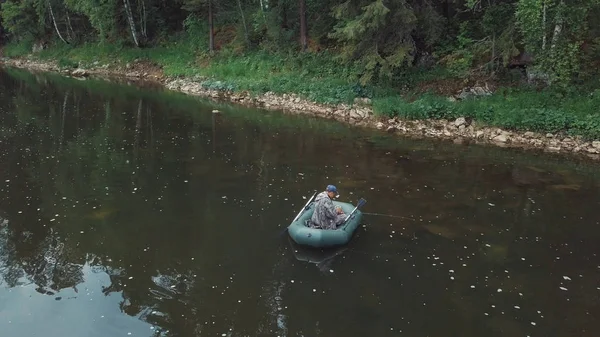 Vista aérea do homem em coverall pesca no barco de borracha no rio perto do lado com plantas, arbustos e árvores no dia de verão. Imagens de stock. Estação de pesca de verão — Fotografia de Stock