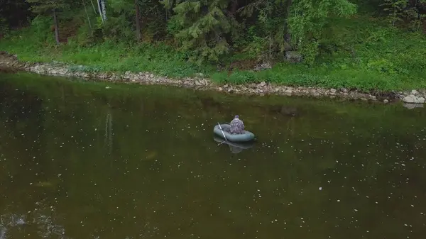 Vista aérea del hombre en mono pescando en el bote de goma en el río cerca del lado con plantas, arbustos y árboles en el día de verano. Imágenes de archivo. Temporada de pesca de verano —  Fotos de Stock