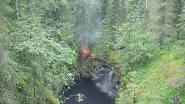 Veduta aerea dell'uomo in piedi sul bordo di un burrone profondo con un bagliore di segnale rosso in mano nella foresta vicino agli alti vecchi alberi e arbusti. Filmati delle scorte. Segnali SOS nella foresta — Foto Stock