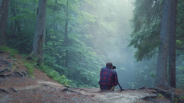 Vista do homem de camisa xadrez e calças sentadas na borda da ravina profunda e tirar uma foto ou vídeo da bela paisagem florestal em um nevoeiro. Imagens de stock. Vista incrível da floresta misteriosa — Fotografia de Stock