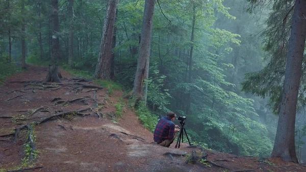 Vista do homem de camisa xadrez e calças sentadas na borda da ravina profunda e tirar uma foto ou vídeo da bela paisagem florestal em um nevoeiro. Imagens de stock. Vista incrível da floresta misteriosa — Fotografia de Stock