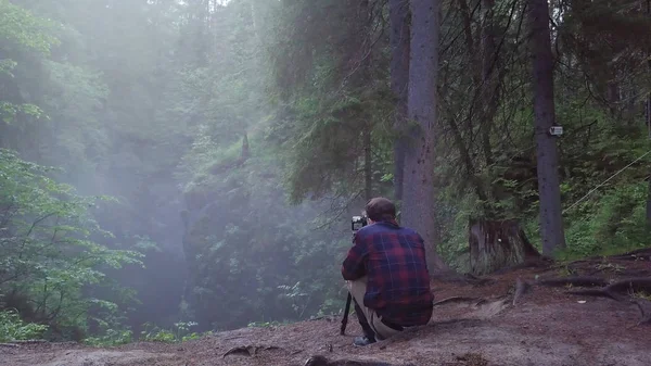 Homem de camisa xadrez e calças sentadas na borda da ravina profunda e tirando uma foto ou vídeo da paisagem em um nevoeiro. Imagens de stock. Vista incrível da floresta misteriosa — Fotografia de Stock
