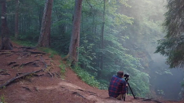 Homem de camisa xadrez e calças sentadas na borda da ravina profunda e tirando uma foto ou vídeo da paisagem em um nevoeiro. Imagens de stock. Vista incrível da floresta misteriosa — Fotografia de Stock