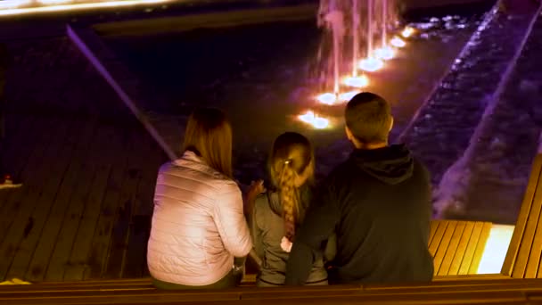 Νεαρή οικογένεια που κοιτάζει το βραδινό σιντριβάνι. Υλικό από μετοχές. Σε όμορφο βράδυ νέα οικογένεια με το παιδί ματιά στα σιντριβάνια της πόλης με τα φώτα — Αρχείο Βίντεο