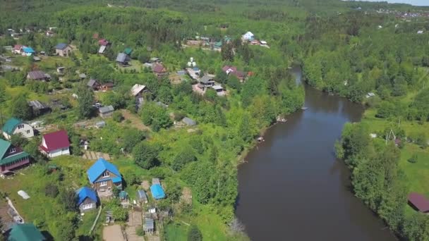 Vista aérea del pueblo y casas cerca del gran río rodeado de coníferas y árboles de hoja caduca en el cálido día de verano. Clip. Hermosa vista desde arriba del pintoresco paisaje rural — Vídeos de Stock