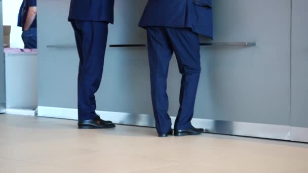 Крупный план двух бизнесменов в синих брюках, пиджаках и черных туфлях, стоящих у стойки регистрации и спрашивающих о чем-то. СМИ. Концепция бизнеса и управления — стоковое видео
