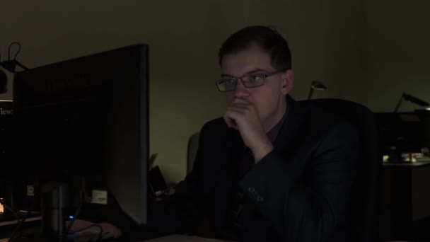 Уставший молодой человек, думающий и выглядящий уставшим в темном офисе на мониторе. Запись. Уставший бизнесмен в очках и работает допоздна за компьютером в офисе . — стоковое видео