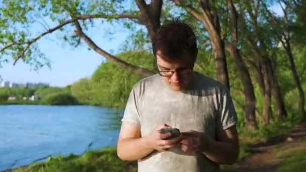 Mladý muž, závislý na telefonu, kráčel lesem podél řeky a díval se na své zařízení. Záběry ze skladu. Chlap, co si píše nebo si něco posílá v letním lese. — Stock video