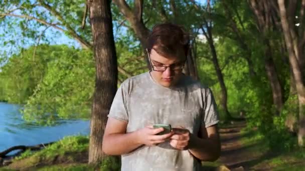 Joven con un móvil en sus manos enviando mensajes de texto a algo durante su aventura en el bosque. Imágenes de archivo. Hombre ignorando la hermosa naturaleza, bosque verde y lago azul, adicción al teléfono . — Vídeo de stock