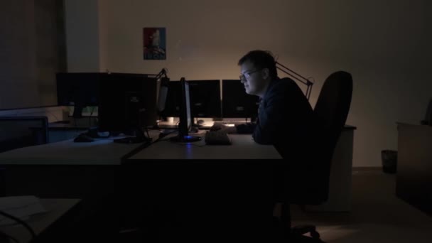 Hardwerkende jonge zakenman aan zijn bureau op zoek naar de informatie op het scherm van zijn computer, zijaanzicht. Voorraadbeelden. Avond op kantoor, laat aan het werk. — Stockvideo