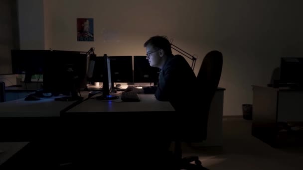 Trabajador joven empresario en su escritorio en busca de la información en la pantalla de su ordenador, vista lateral. Imágenes de archivo. Noche en la oficina, trabajando hasta tarde . — Vídeo de stock