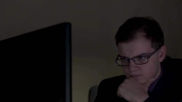 Joven hombre de negocios con gafas trabajando hasta tarde en la oficina, tocándose la barbilla y luciendo serio. Imágenes de archivo. Joven trabajando en la computadora por la noche en la oficina oscura . — Vídeos de Stock