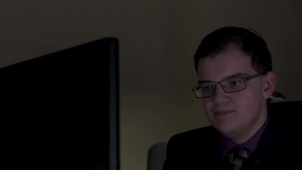 Joven hombre de negocios con gafas trabajando hasta tarde en la oficina y sonriendo con una pared en el fondo. Imágenes de archivo. Joven trabajando en la computadora por la noche en la oficina oscura . — Vídeos de Stock