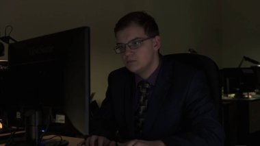 Yorgun genç adam düşünme ve monitörde karanlık ofiste yorgun görünümlü. Stok görüntüleri. Yorgun işadamı gözlük takıyor, kahve içiyor, ofiste bilgisayarda geç saatlere kadar çalışıyor.