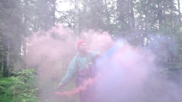 Hombre con bombas de humo de colores en la naturaleza. Imágenes de archivo. Joven hombre extiende corrientes de coloridas damas de humo en bosque denso — Vídeo de stock