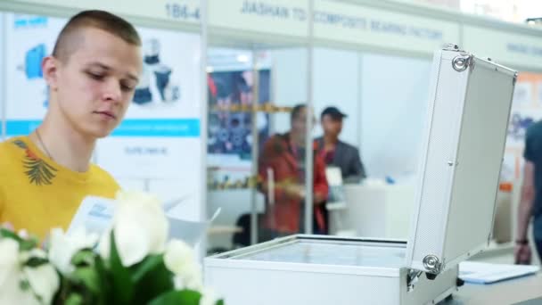 Närbild av ung man i gul t-shirt stående med vissa dokument och knacka på skärmen på datorn i silver resväska. Media. Robotics forum utställning — Stockvideo