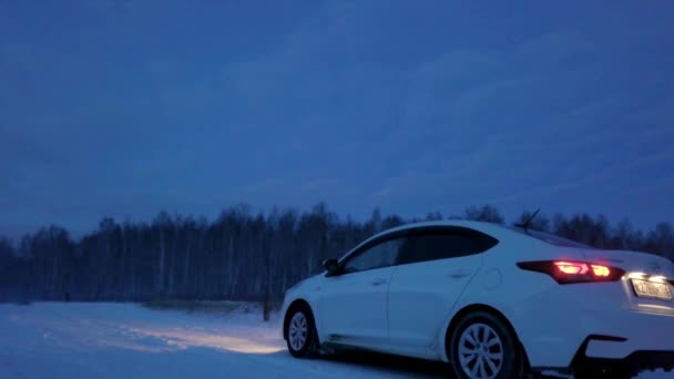 Samochód na zimowej drodze w pobliżu lasu w godzinach wieczornych. Materiały stockowe. Widok z tyłu samochodu na pokrytej śniegiem drodze w pobliżu lasu pasa późnym wieczorem — Wideo stockowe