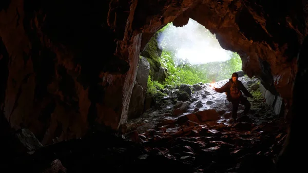 Wandelaar naar beneden in grot. Stock footage. Reiziger met rode signaal kaars daalt in grot op helling op achtergrond van gewelfde ingang — Stockfoto