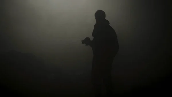 Black silhouette of man standing in smoky dark. Stock footage. Mysterious silhouette of young man stands in darkness lit only by dim light in haze