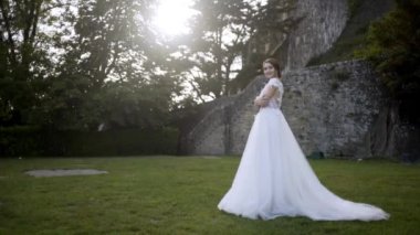 Ortaçağ duvarlarının arka planda güzel gelin. Eylem. Ağaçların arka planda yeşil çim üzerinde poz beyaz elbiseli Gelin. Gelin yeşil bahçede gelinlik şefkatle gülümsüyor