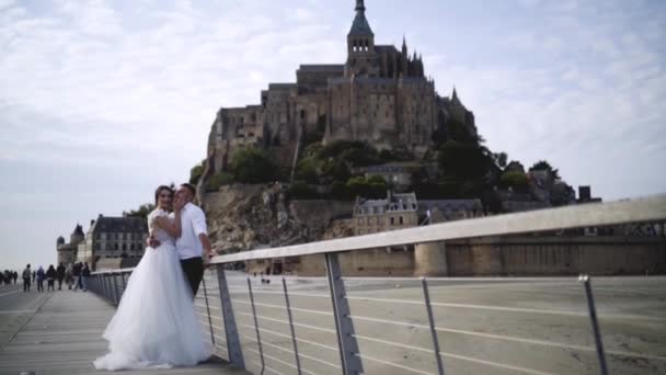 Jonggehuwden op achtergrond van oude kasteel op heuvel op achtergrond van de hemel. Actie. Mooie jonge paar in trouwjurken staan op achtergrond van middeleeuws kasteel — Stockvideo