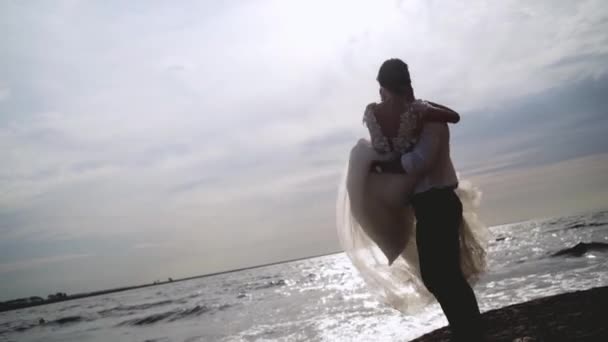 Bruidegom houdt bruid in zijn armen op de achtergrond van de zee. Actie. Bruidegom opgepikt bruid en draait haar in de zon op achtergrond van zee landschap. Oprechte en stralende liefde van jonggehuwden — Stockvideo