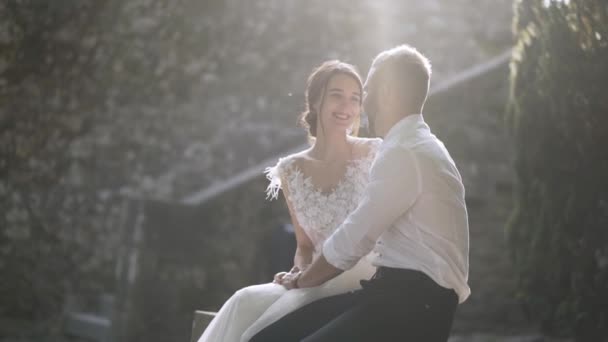 Belos recém-casados sentados à luz do sol. Acção. Bela noiva e noivo sentar e olhar um para o outro iluminado pela luz solar brilhante. Amor sincero e terno pelos jovens — Vídeo de Stock