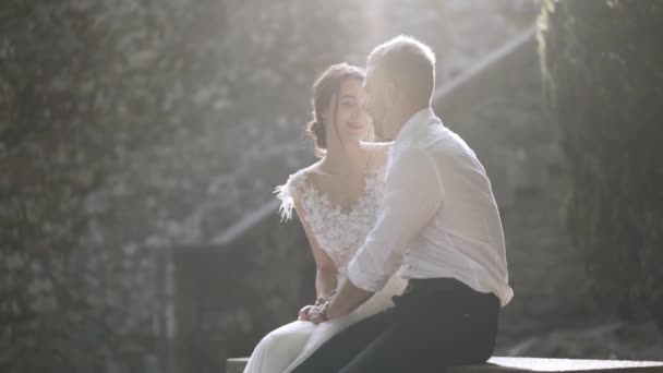 Beautiful newlyweds sitting in sunlight. Action. Beautiful bride and groom sit and look at each other illuminated by bright sunlight. Sincere and tender love of young people — Stock Video