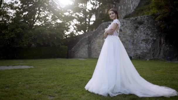Beautiful bride on background of medieval walls. Action. Bride in white dress posing on green lawn on background of trees. Bride smiles tenderly in her wedding dress in green garden — Stock Video