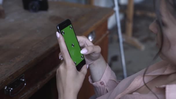 Blick von der Schulter einer jungen Frau in rosafarbenem Hemd mit dunklen Haaren, die auf ihrem Smartphone mit grünem Bildschirm tippt. Archivmaterial. Smartphone neues Technologie-Konzept — Stockvideo