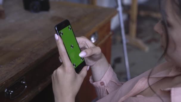 Mujer joven en camisa rosa con el pelo oscuro sentado en la mesa y e en su teléfono inteligente con pantalla verde croma clave. Imágenes de archivo. Pantalla de clave de croma para la colocación de su propio contenido . — Vídeo de stock