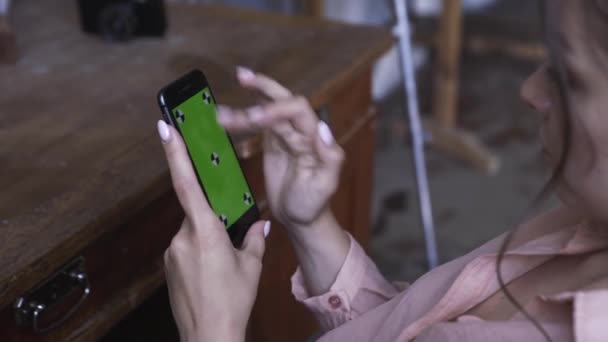 Mujer joven en camisa rosa con el pelo oscuro sentado en la mesa y e en su teléfono inteligente con pantalla verde croma clave. Imágenes de archivo. Pantalla de clave de croma para la colocación de su propio contenido . — Vídeos de Stock