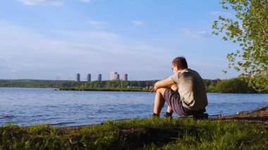Genç adam şehrin arka planda nehir kıyısında oturur. Stok görüntüleri. İnsan nehir kıyısında oturan doğa manzaralarını sever. Doğa, nehir ve arka planda dalgalar şehrin yüksek katlı görebilirsiniz