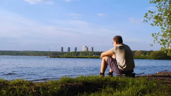 Genç adam şehrin arka planda nehir kıyısında oturur. Stok görüntüleri. İnsan nehir kıyısında oturan doğa manzaralarını sever. Doğa, nehir ve arka planda dalgalar şehrin yüksek katlı görebilirsiniz