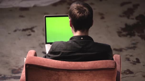 Uitzicht vanaf de achterkant van de man in zwart shirt zittend op de bruine stoel en typen op laptop met groen Chroma Key-scherm. Stock footage. Laptop nieuw technologie concept, Chroma Key groen scherm — Stockvideo