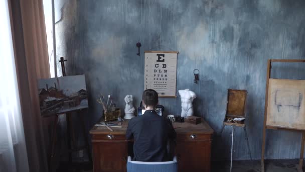 Fiatal férfi fekete inget ül az asztalnál, kézműves területen, és dolgozik az ő laptop. Stock Footage. Szabadúszóként otthon, Kilátás vissza — Stock videók