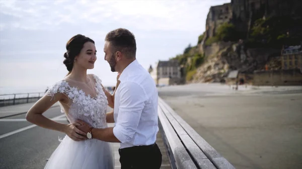 Pár a vízparton a sziklák. Akció. Newlyweds átölelve a vízparton a háttérben a sziklák és az ég. Menyasszony és vőlegény ölelés a háttérben a gyönyörű táj rakparton — Stock Fotó