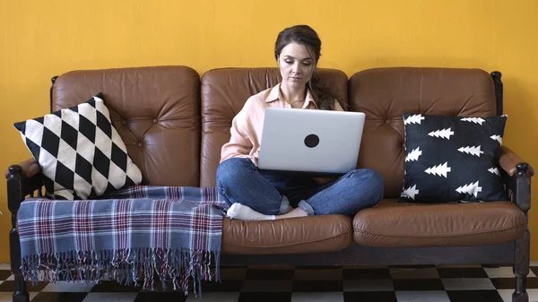 Κοντινό πλαίσιο της νεαρής χαμογελαστή γυναίκα σε ροζ πουκάμισο και Τζιν κάθονται στον καναπέ και εργάζονται ή σέρφινγκ στο διαδίκτυο σχετικά με το φορητό υπολογιστή. Υλικό από μετοχές. Ελεύθερος επαγγελματίας στο σπίτι — Φωτογραφία Αρχείου