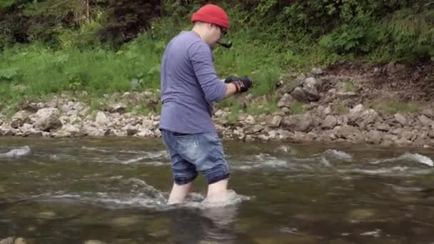 O homem tira fotos da água do rio. Imagens de stock. Fotógrafo profissional tira fotos do fluxo do rio para revista de viagens. Homem fica de joelhos na água e tirar fotos da natureza — Vídeo de Stock